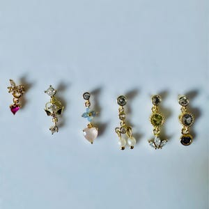 Puede incluir: Seis pendientes dorados con varios diseños. Los pendientes presentan piedras preciosas en rosa, transparente, azul y verde, junto con perlas. Los diseños incluyen corazones, flores y formas geométricas. Se muestran sobre una superficie blanca.