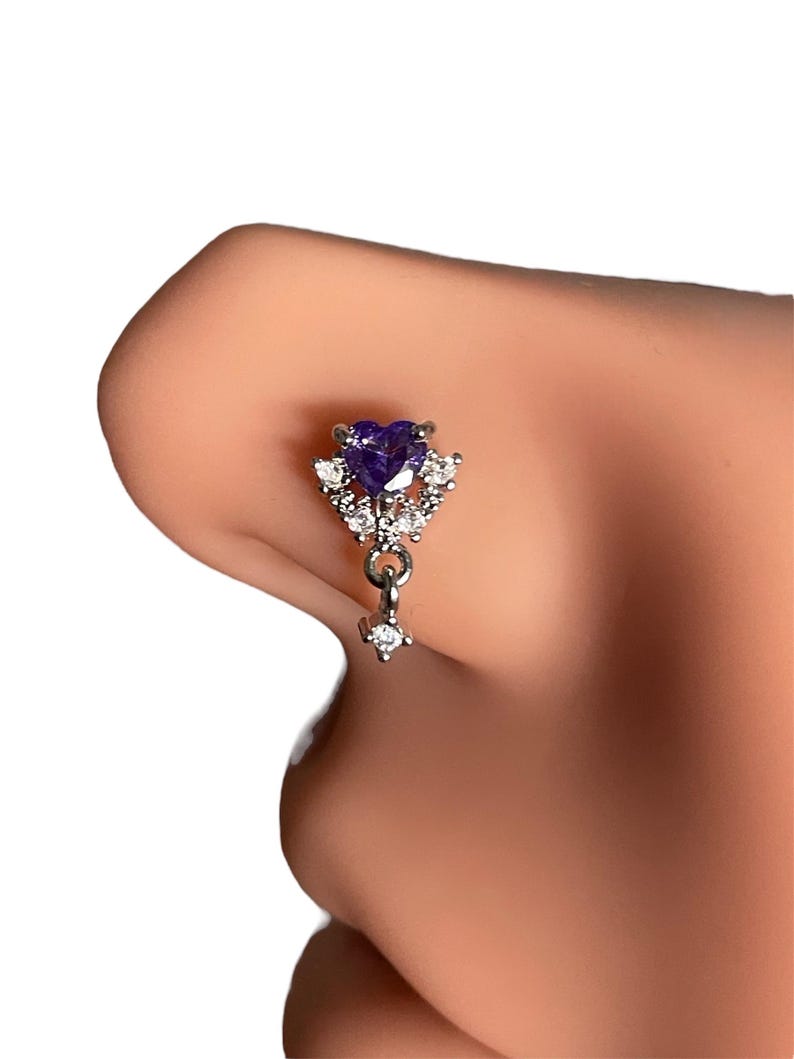 May include: A silver nose ring with a dangling charm. The charm features a purple heart-shaped stone surrounded by smaller clear stones.