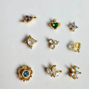 May include: A collection of nine gold-toned stud earrings featuring various shapes and gemstones. Designs include a purple heart with an arrow, a green heart with wings, a flower, a circular clear stone, a three-stone cluster, a heart outline, and floral motifs.