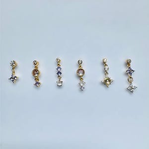 Puede incluir: Seis pendientes colgantes dorados con varios diseños de piedras preciosas. Cada pendiente presenta un pequeño perno redondo con una disposición colgante de piedras preciosas claras, moradas y rosas. Los pendientes se muestran sobre un fondo blanco liso.