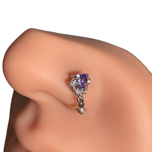 May include: A silver nose ring with a dangling charm. The charm is shaped like a key with a purple gemstone at the top and clear rhinestones around it.