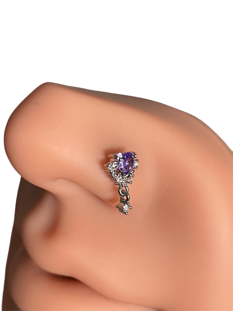 May include: A silver nose ring with a dangling charm. The charm is shaped like a key with a purple gemstone at the top and clear rhinestones around it.