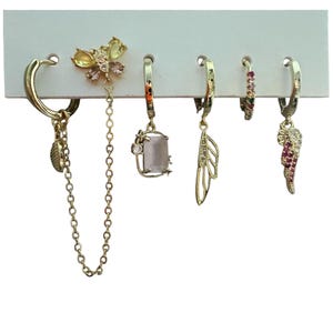 Puede incluir: Un conjunto de cinco pendientes de aro dorados presentados sobre una tarjeta blanca. El set incluye un dije de mariposa con una cadena larga, un colgante de piedra rectangular, un dije de ala, un aro pequeño con piedras de colores y un ala rosa.