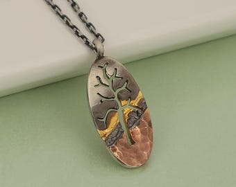 May include: Silver-tone necklace with an oval pendant featuring a tree design. The pendant has a textured copper and gold-tone inlay. The chain is a dark gray color, and the background is a light green.