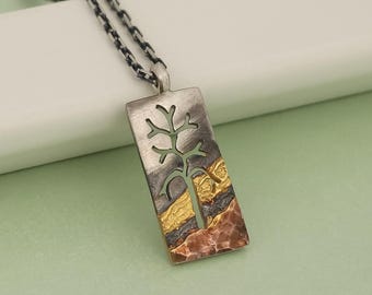 May include: Silver pendant necklace with a rectangular pendant featuring a cut-out tree design. The pendant has a textured, mixed-metal appearance with gold and copper accents. The chain is a dark, linked style.