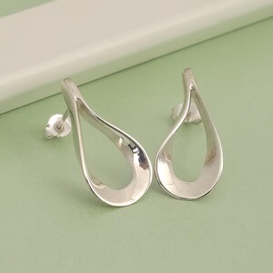 Pode incluir: Um par de brincos de prata em forma de gota. Os brincos têm um acabamento liso e polido e um centro oco. Estão colocados sobre um fundo verde claro.