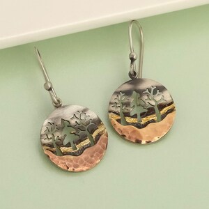 Può includere: Un paio di orecchini pendenti in argento con un design circolare. Ogni orecchino presenta un paesaggio con alberi ritagliati nel metallo, con un fondo color rame e una parte superiore argentata. Gli orecchini pendono da ganci d'argento.