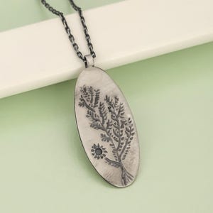 May include: A silver-toned pendant necklace featuring an oval pendant with an etched tree and sun design. The pendant hangs from a dark chain. The background is a light green color.