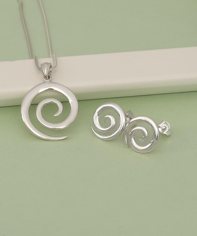 May include: A silver necklace and earrings set featuring a spiral design. The necklace has a chain and a pendant with a single spiral. The earrings have two spirals each.