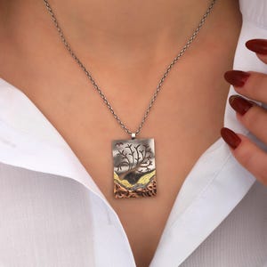 Puede incluir: Un collar de cadena de plata con un colgante rectangular que presenta un diseño de árbol. El colgante está hecho de metales mixtos, incluyendo plata, cobre y oro.