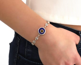 May include: A silver chain bracelet with a blue and yellow evil eye charm. The charm is round and has a white outline.