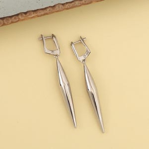 May include: A pair of silver drop earrings. Each earring has a rectangular top with a hinged closure and a long, pointed, teardrop-shaped pendant. The earrings are set against a light yellow background.