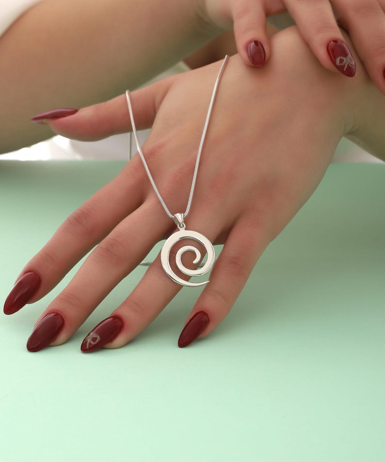May include: A silver necklace with a spiral pendant. The pendant is a circle with a spiral design inside. The necklace is on a woman's hand with red nail polish.