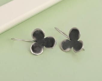 May include: A pair of silver-toned earrings. Each earring features a three-leaf clover design with a textured surface. The earrings have a simple hook closure. The background is a light green color.