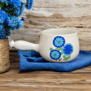 May include: White ceramic jug with a blue and green floral design. The jug has a handle and is sitting on a blue cloth.