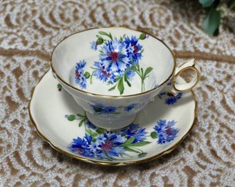May include: A white teacup and saucer set with a floral design. The teacup and saucer are decorated with blue flowers and green leaves. The rims are trimmed with gold. The teacup has a curved handle.