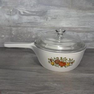 May include: White ceramic saucepan with a glass lid. The saucepan has a long handle and a decorative pattern of colorful vegetables, including tomatoes and lemons. A vintage kitchenware item.