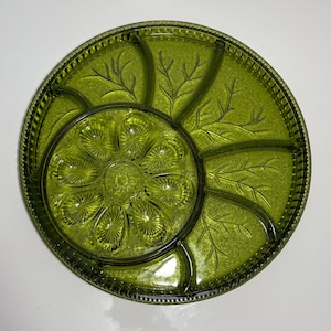 May include: A round, olive green glass serving dish with a central floral design and six surrounding compartments. The compartments feature a leaf pattern. The rim has a textured, beaded design. This vintage dish is perfect for serving snacks or appetizers.