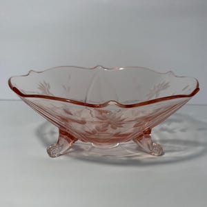 May include: A pink glass serving bowl with a scalloped rim and three decorative feet. The bowl features etched floral designs and a flared, angled shape. The glass has a translucent quality, and the bowl is set against a white background.