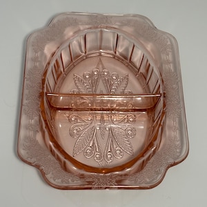May include: A vintage pink glass butter dish with a decorative floral and geometric design. The dish has a rectangular base with a raised, oval-shaped interior section. The dish features a detailed embossed pattern and is made of transparent pink glass.