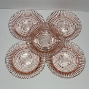 May include: A set of five round, light pink glass plates featuring a pressed radial sunburst pattern in the center and a ribbed, fluted edge design. The translucent glass items are arranged in a cluster, showcasing the consistent circular texture and delicate pink hue across each individual piece of glassware.
