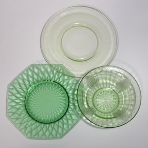 May include: Three vintage glass plates in shades of green and clear. The plates include a clear plate, a green octagonal plate with a diamond pattern, and a green bowl with a sunburst design.