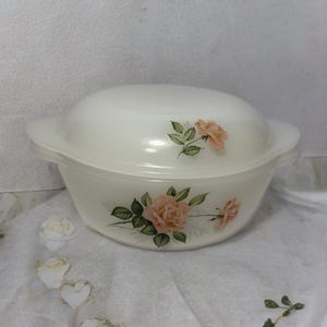 May include: A white, oval-shaped casserole dish with a matching lid. The dish is decorated with pink roses and green leaves. The dish has two handles and is sitting on a white surface.