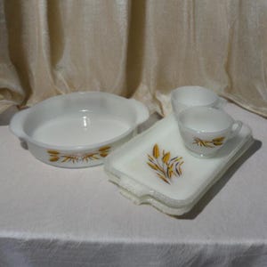 May include: A set of vintage white milk glass serving dishes with a wheat stalk design. Includes a round casserole dish, rectangular trays, and a teacup and saucer. The wheat design features gold and brown colours.