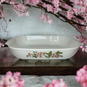 May include: A rectangular, off-white ceramic baking dish with rounded corners and small handles. The dish features a floral design with pink blossoms and green leaves. The background includes pink flowers.