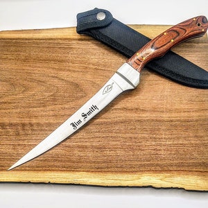 May include: A personalized, silver-colored,  fillet knife with a wooden handle. The knife is engraved with the name "Jim Smith" and is resting on a wooden cutting board. The knife is accompanied by a black nylon sheath.