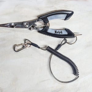 May include: Fishing pliers with a black and silver design, featuring the name "Ralph" in a stylized font. Includes a coiled lanyard with a carabiner clip for secure attachment. The pliers are made of stainless steel.