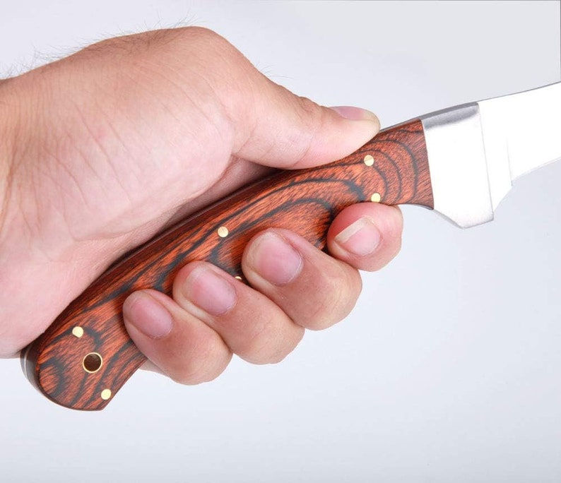 May include: A close-up of a person's hand holding a hunting knife with a wooden handle. The handle is a dark red wood with gold accents. The blade is silver and has a slight curve.
