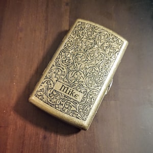 May include: A gold metal cigarette case with an engraved floral design and the name "Mike" on the front.