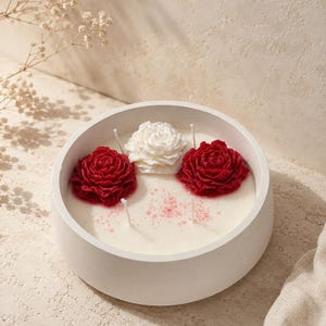 Puede incluir: Una vela redonda blanca con tres mechas y tres adornos florales. Dos velas rojas en forma de rosa flanquean una vela blanca en forma de rosa. La vela está en un recipiente redondo blanco. El fondo es de color beige neutro.