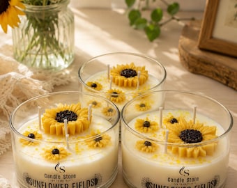 May include: Three glass jar candles with sunflower designs. Each candle has a large sunflower and several smaller ones on top of the creamy wax. The text "SUNFLOWER FIELDS" is printed on the front of the jars. A vase of sunflowers is in the background.