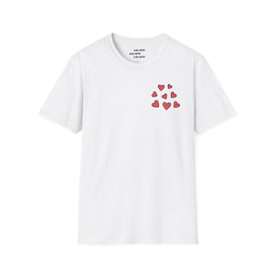 Puede incluir: Camiseta blanca con un diseño de corazones rojos en la parte delantera. Los corazones están dispuestos en un grupo y están delineados en negro. El texto "LUVVV" está impreso en el cuello de la camiseta.