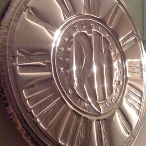 May include: A circular, silver-colored clock face with Roman numerals and the words "Time Block Company" inscribed around the center.