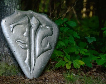 May include: A silver metal shield with a raised design of two stylized animal heads and a sword. The shield is leaning against a tree trunk in a forest setting.