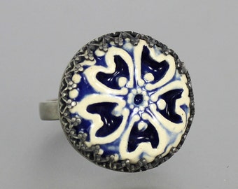 May include: A silver ring with a blue and white ceramic flower design. The flower has a white center with a blue outline and four petals.