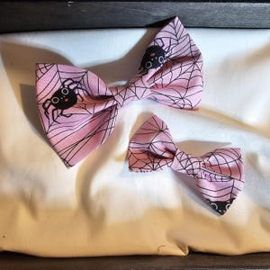 May include: Two bow ties in a light pink fabric featuring a black spider web pattern and small black spiders with white eyes. The larger bow tie is positioned above the smaller bow tie, both displaying the same repeating graphic print design.
