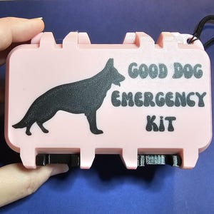 May include: A light pink rectangular emergency kit case featuring a black silhouette of a German Shepherd dog and the black text "GOOD DOG EMERGENCY KIT" printed on the front. The case has a textured finish and black latch closures.