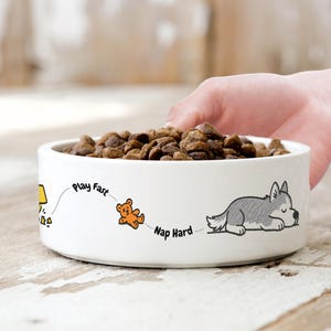 May include: A white ceramic pet bowl filled with brown kibble, featuring a graphic illustration of a grey husky sleeping next to an orange teddy bear, with the text "Play Fast" and "Nap Hard" printed in black along the side of the bowl.