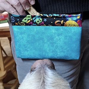 以下が含まれることがあります： ターコイズと黒の犬用おやつポーチ。カラフルな誕生日のテーマ柄。ポーチは人が持ち、犬が見上げています。犬のおやつが見えます。