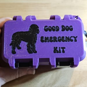 Op de afbeelding: Een rechthoekige paarse plastic opbergdoos met een zwart silhouet van een hond en de zwarte tekst "GOOD DOG EMERGENCY KIT" op de voorzijde. De doos heeft een getextureerd oppervlak, een scharnierend deksel en een zwarte riem aan de zijkant.