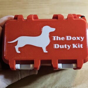 May include: A rectangular orange plastic case with a white silhouette of a dachshund and the white text "The Doxy Duty Kit" printed on the front. The case features a hinged lid with white latches and a black wrist strap attached to the side.
