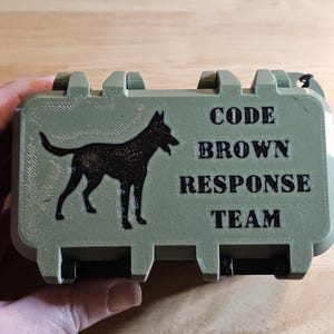 May include: An olive green rectangular storage case with a black silhouette of a dog on the left side and the black text "CODE BROWN RESPONSE TEAM" printed on the right side. The case features a rugged design with visible hinges and a black strap.