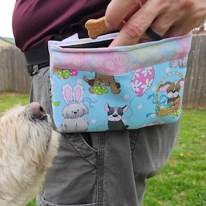 以下が含まれることがあります： 子犬、イースターエッグ、風船が描かれた、イースターをテーマにしたカラフルなプリントが施された、水色の犬用おやつバッグ。バッグは人が持っており、犬がバッグを見ています。バッグには黒いストラップが付いており、骨の形をしたおやつが見えます。