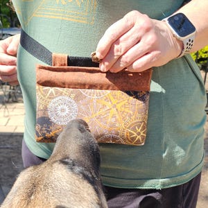 以下が含まれることがあります： 茶色とタン色の犬用おやつポーチ。コンパスローズのデザインで、ウエストに巻かれた黒いストラップに取り付けられています。ポーチは人が開けており、犬がポーチを見上げています。