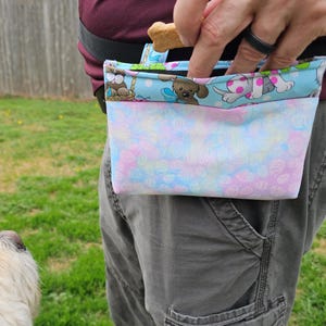 以下が含まれることがあります： 犬のおやつポーチ。水色のトップには漫画の犬が描かれ、パステルカラーのボトムには卵の模様があります。ポーチは黒いベルトに取り付けられ、ウエストに着用します。犬のおやつがポーチの上に持たれています。