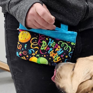 May include: A black and turquoise dog treat bag with a colorful "Happy Birthday" design. The bag features balloons, confetti, and stars. The bag is held by a person.